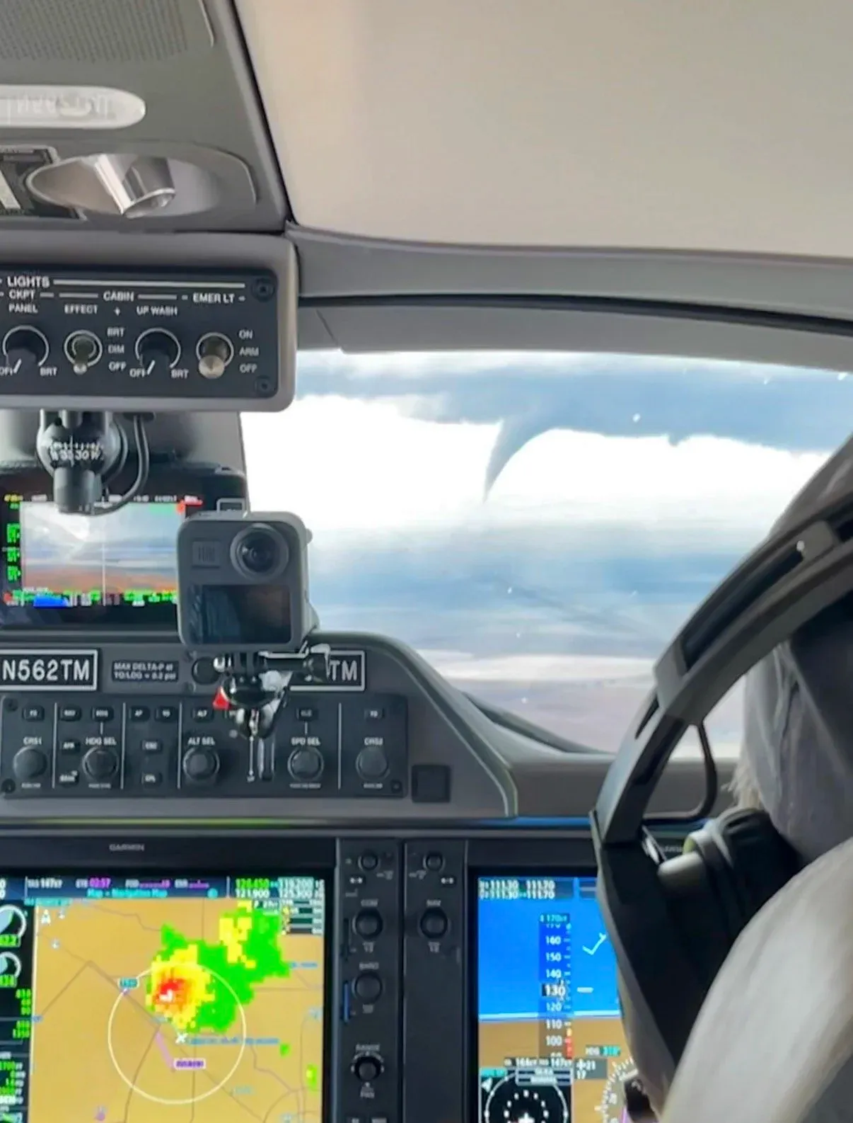 Inside an airplane cockpit: instrument panels and GPS screens showing weather radar with a storm, and a tornado visible in the distance through the windshield.