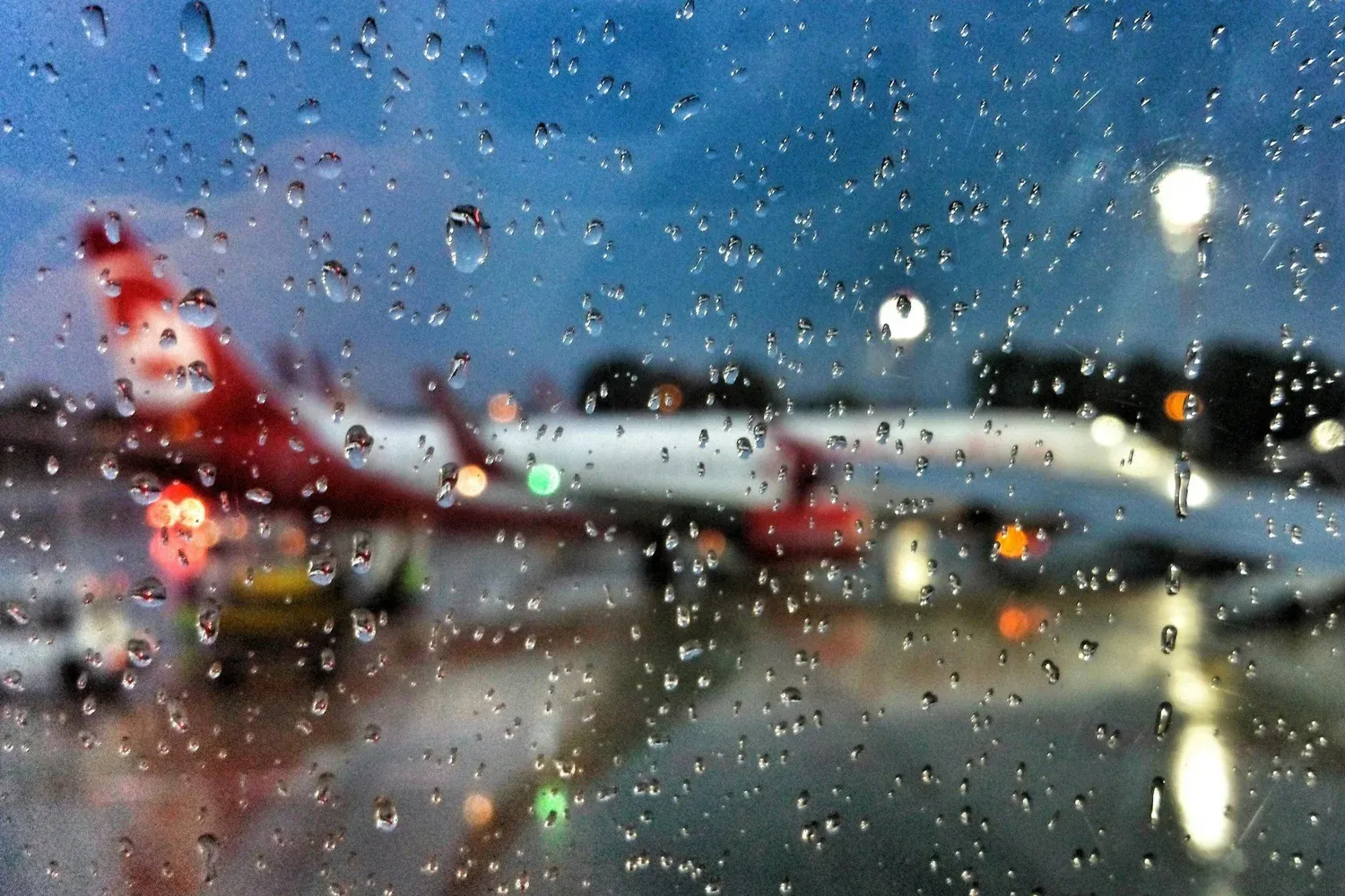 Rain-covered airplane window at night with blurred airport lights.