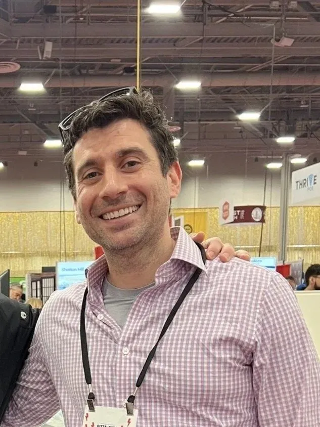 Joey Picca, a smiling man with curly dark hair wearing a pink checkered shirt and a lanyard, standing in a conference hall.