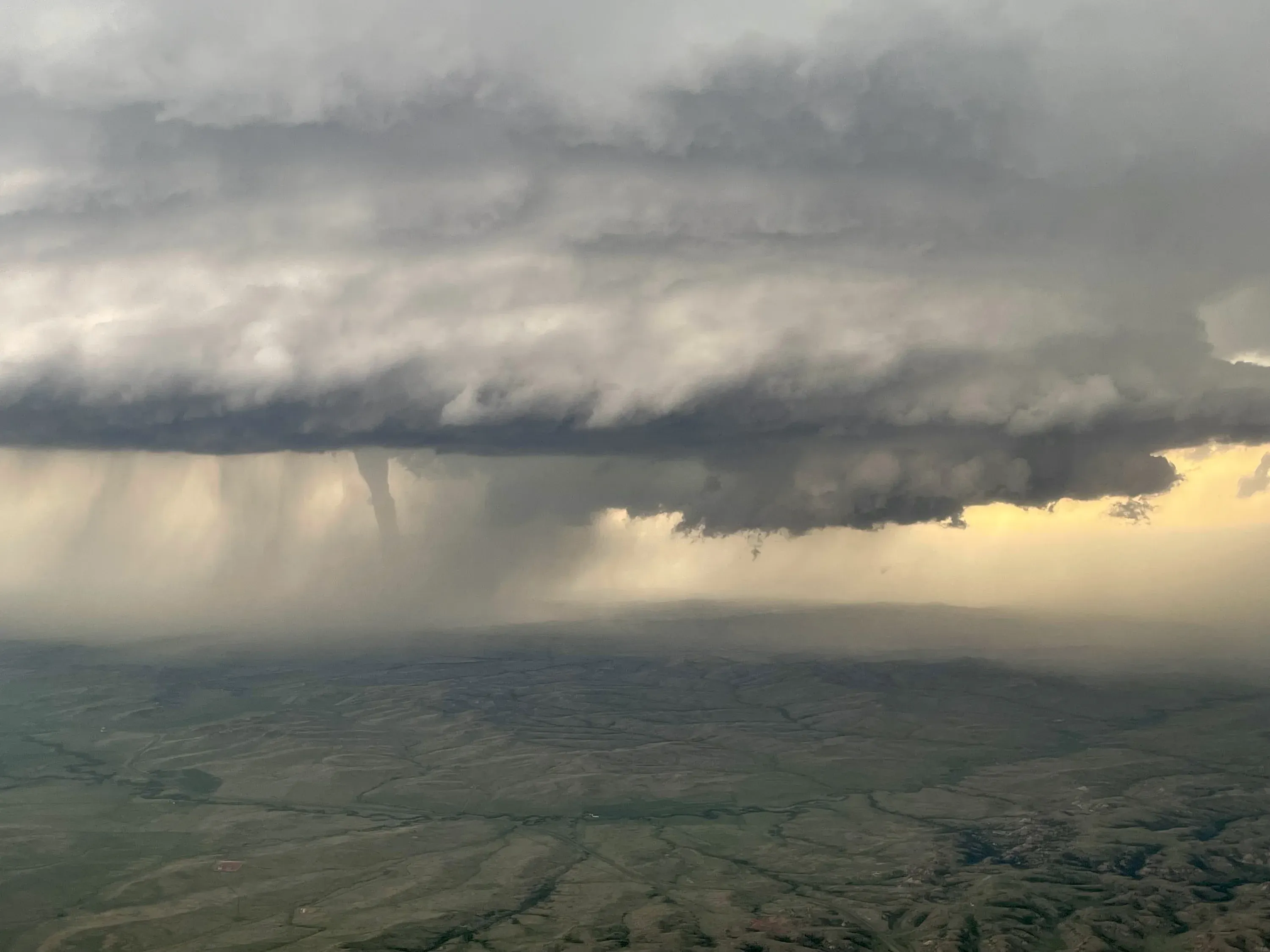 A tornado beneath a dark storm cloud base, seen from the air, with rain and golden light behind it.